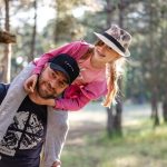 A joyful father giving his daughter a piggyback ride in a sunny forest setting.